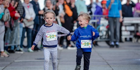 Powiększ grafikę: Dwójka biegnących małych dzieci z przypiętymi numeramin startoywmi, w tle stojący i dpingujący kibice