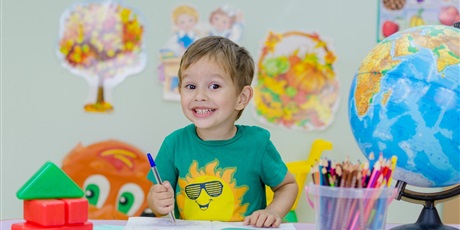 Powiększ grafikę: Uśmiechnięty chłopiec w zielonej koszulce z długopisem w ręku, siedzacy przy stoliku, na którym stoja kredki, globus.